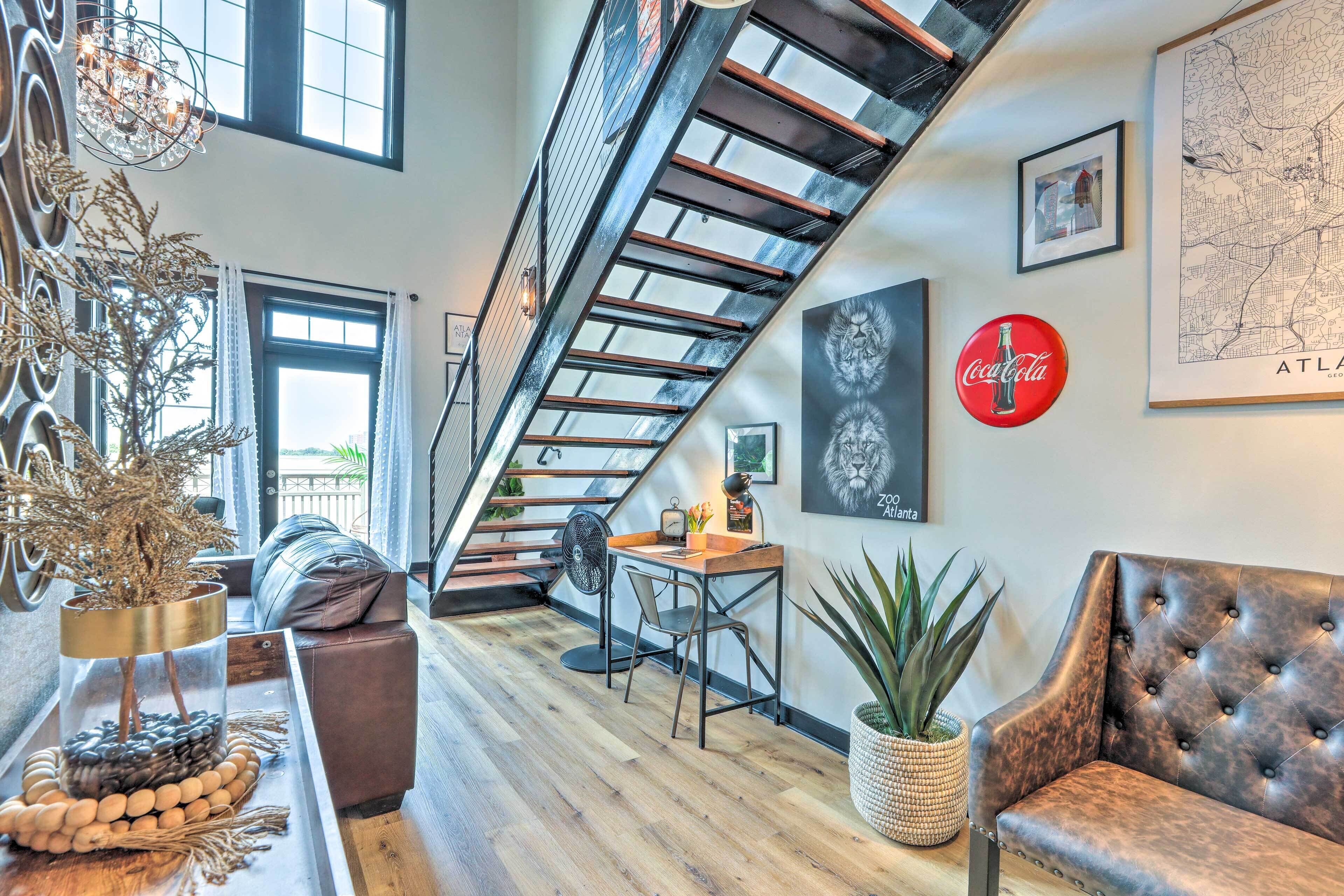 A modern loft with a loft bed, a dining area with a wooden table and chairs, a seating area with a leather couch, and a staircase with a glass railing.