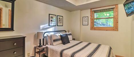 A bedroom with a bed, dresser, lamp, and window with a view of trees.