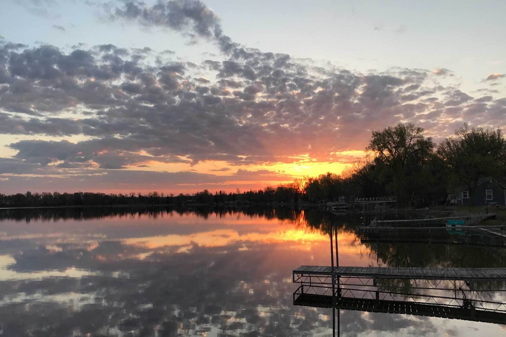 A sunset over a calm lake with a dock and trees in the foreground.