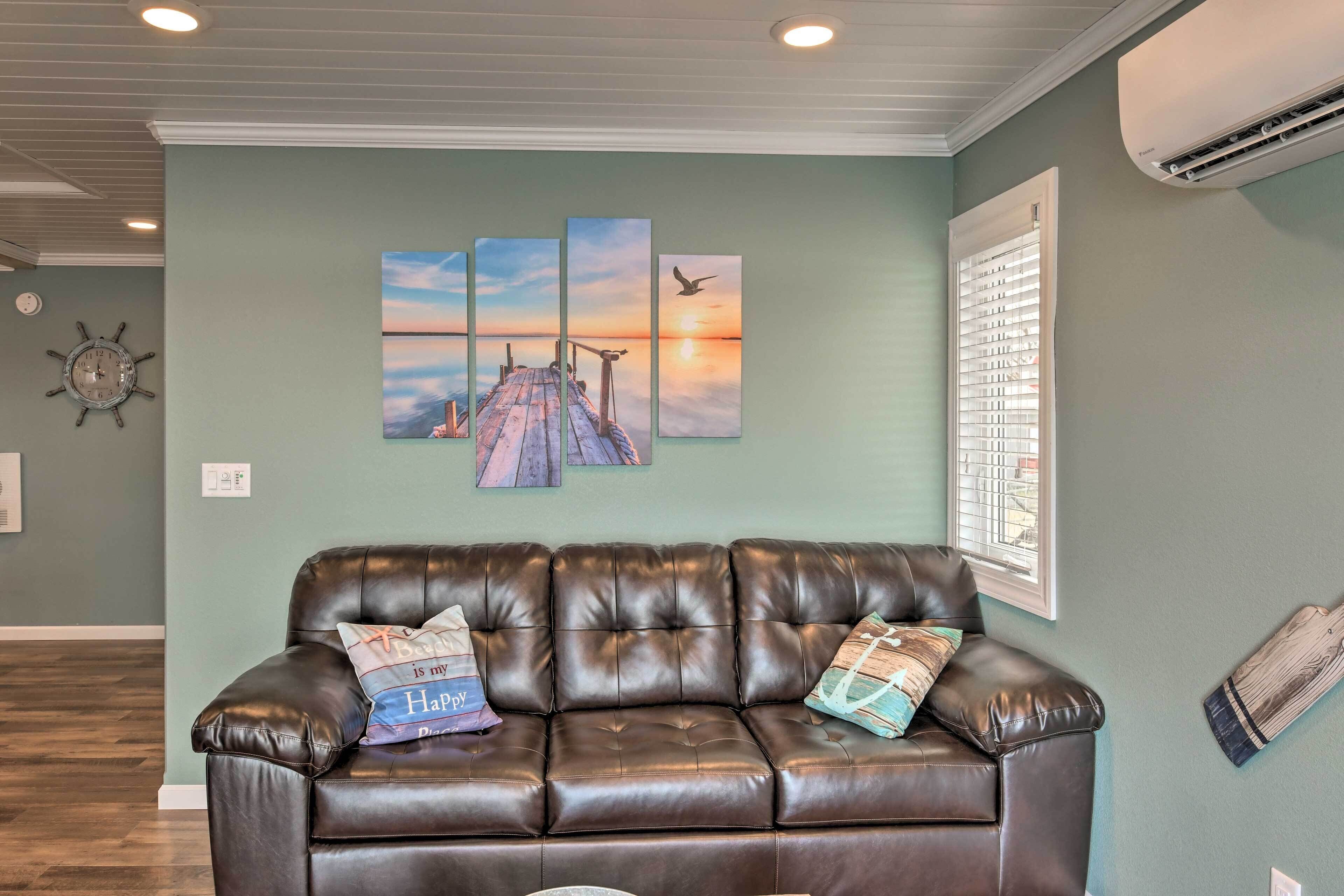 A living room with a brown leather couch, a multi-panel sunset artwork, a clock, and a window with blinds.