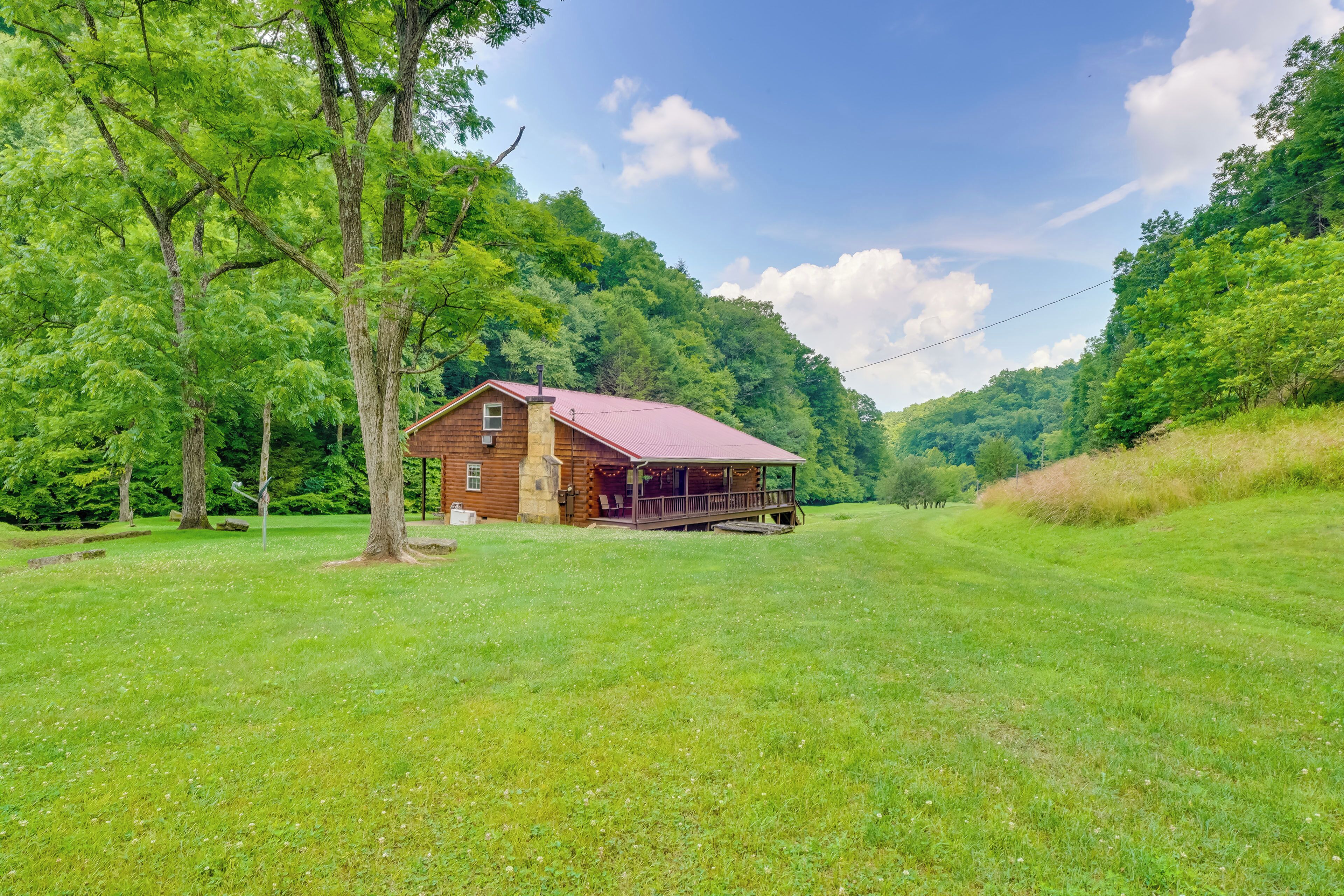 A rustic wooden cabin with a red roof, surrounded by lush green trees and a grassy field.