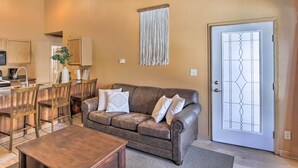 A cozy living area with a brown leather couch, wooden chairs, a coffee table, and a dining table. A decorative wall hanging is above the couch, and a door with a glass panel is visible. - Riverfront Crivitz Condo w/ Fireplace & Views (Crivitz)