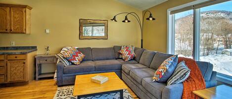 A living room with a gray sectional sofa, a wooden coffee table, and a large window with a view of a snowy landscape.