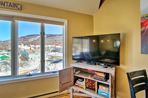 A living room with a large window, a flat-screen TV, and a wooden entertainment unit with books and games. - Lincoln Family Condo: 2 Mi to Loon Mountain Resort (Lincoln)