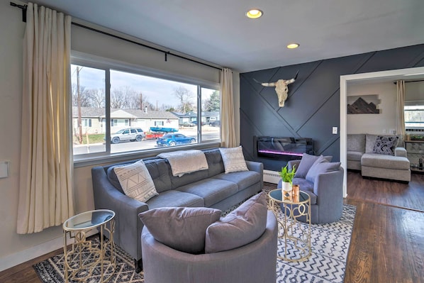 A modern living room with a grey sofa, a round armchair, and a TV. A bull skull decor is on the wall, and there's a window with curtains.