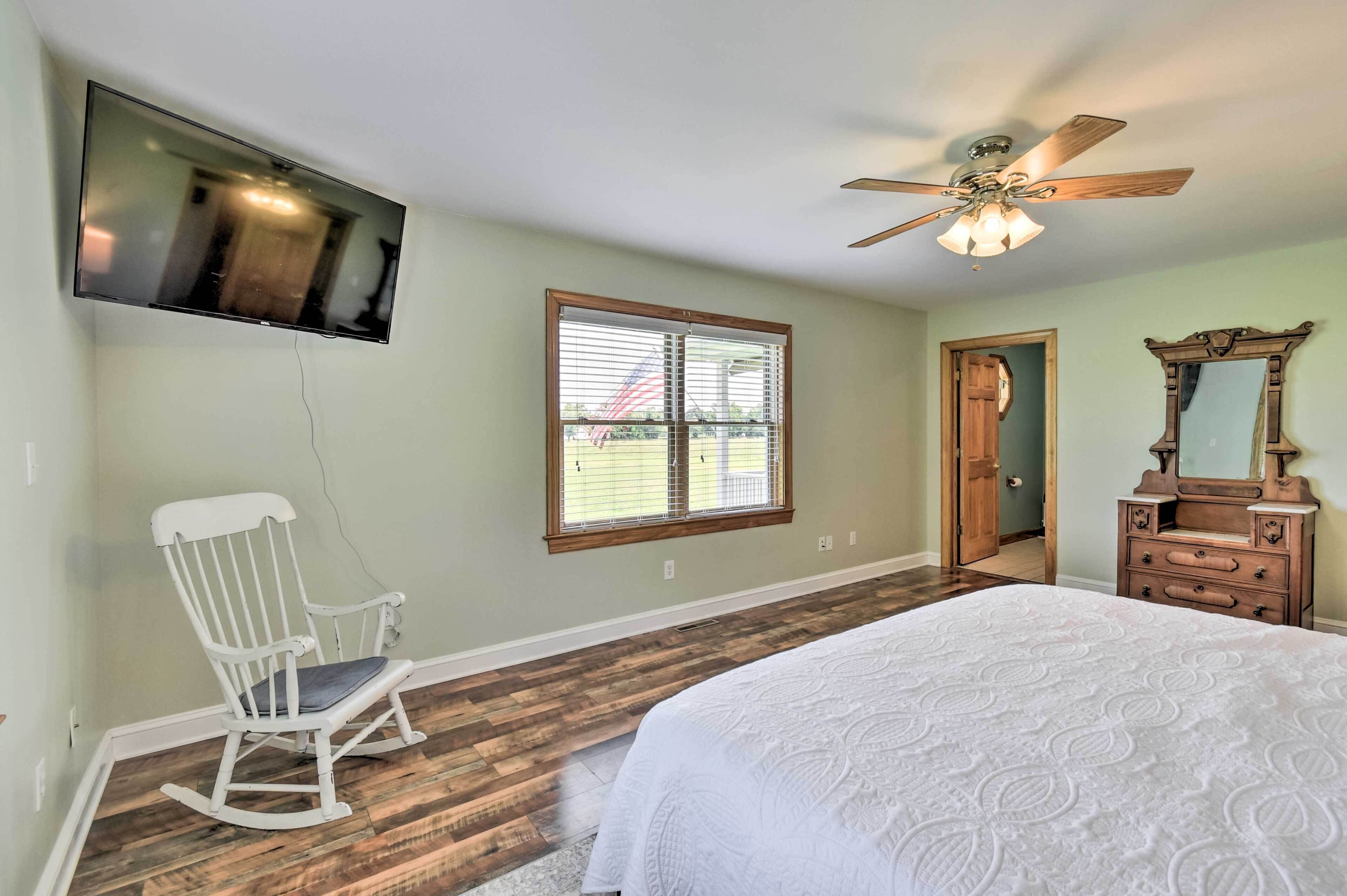 A bedroom with a bed, a rocking chair, a TV mounted on the wall, a window with blinds, a ceiling fan, and a dresser with a mirror.