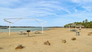 A beach with picnic tables and umbrellas, a trash bin, and scattered rocks.