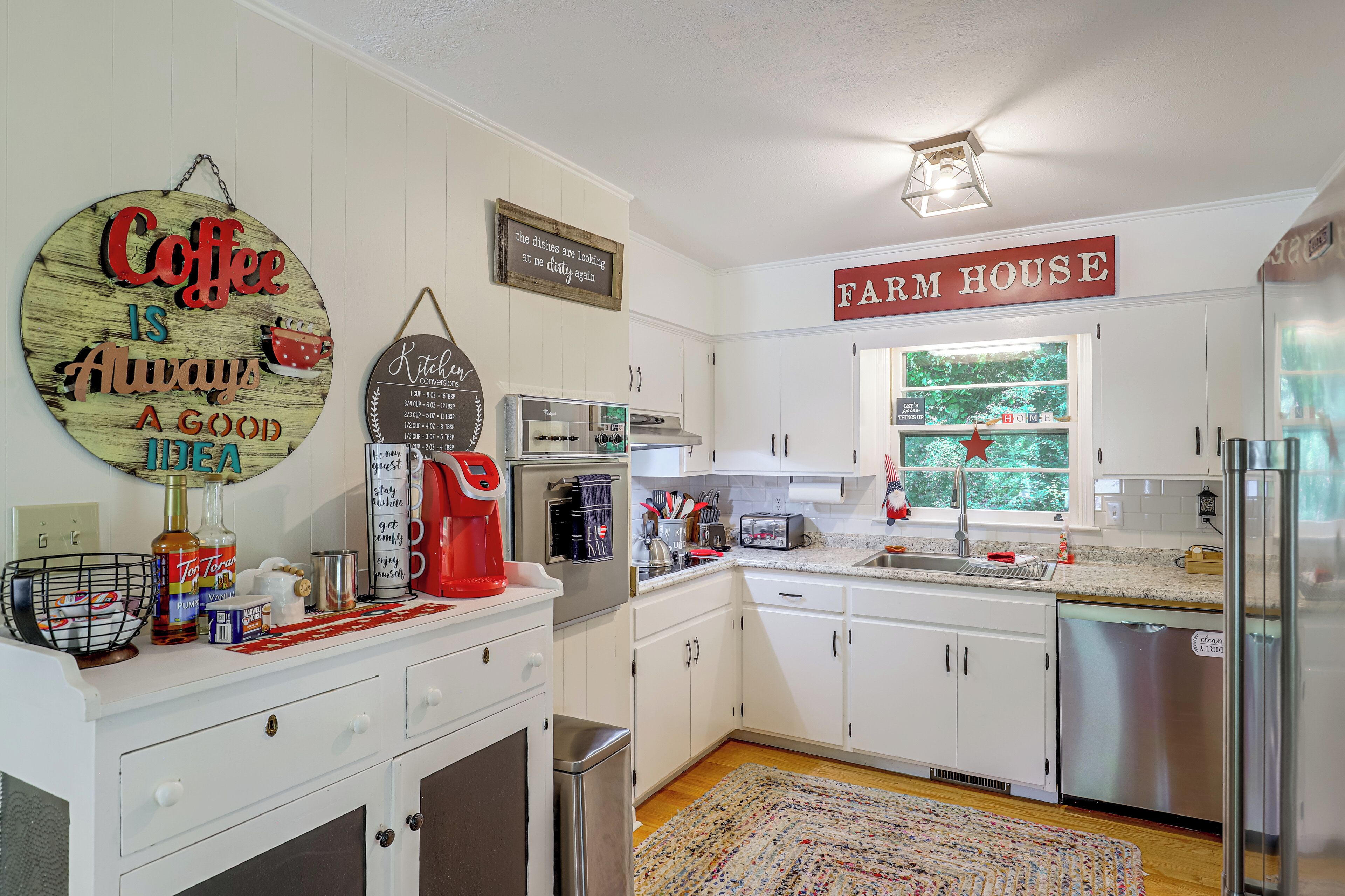 A kitchen with white cabinets, a coffee sign, a farmhouse sign, and a window with a view of greenery.