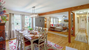A dining area with a wooden table, chairs, and a chandelier. There is a living room in the background with a sofa and a fireplace. - Fire Pit & Game Room: Family Home in Marshall! (Weaverville)