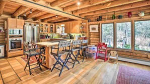 A rustic cabin interior with wooden beams, a kitchen with a refrigerator, stove, and bar area, and large windows overlooking a forest. - Game Room w/ Speakeasy Saloon: Accord Retreat (Accord)