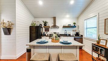A kitchen with white wooden walls, a central island with stools, a stove, and a hanging pot rack.