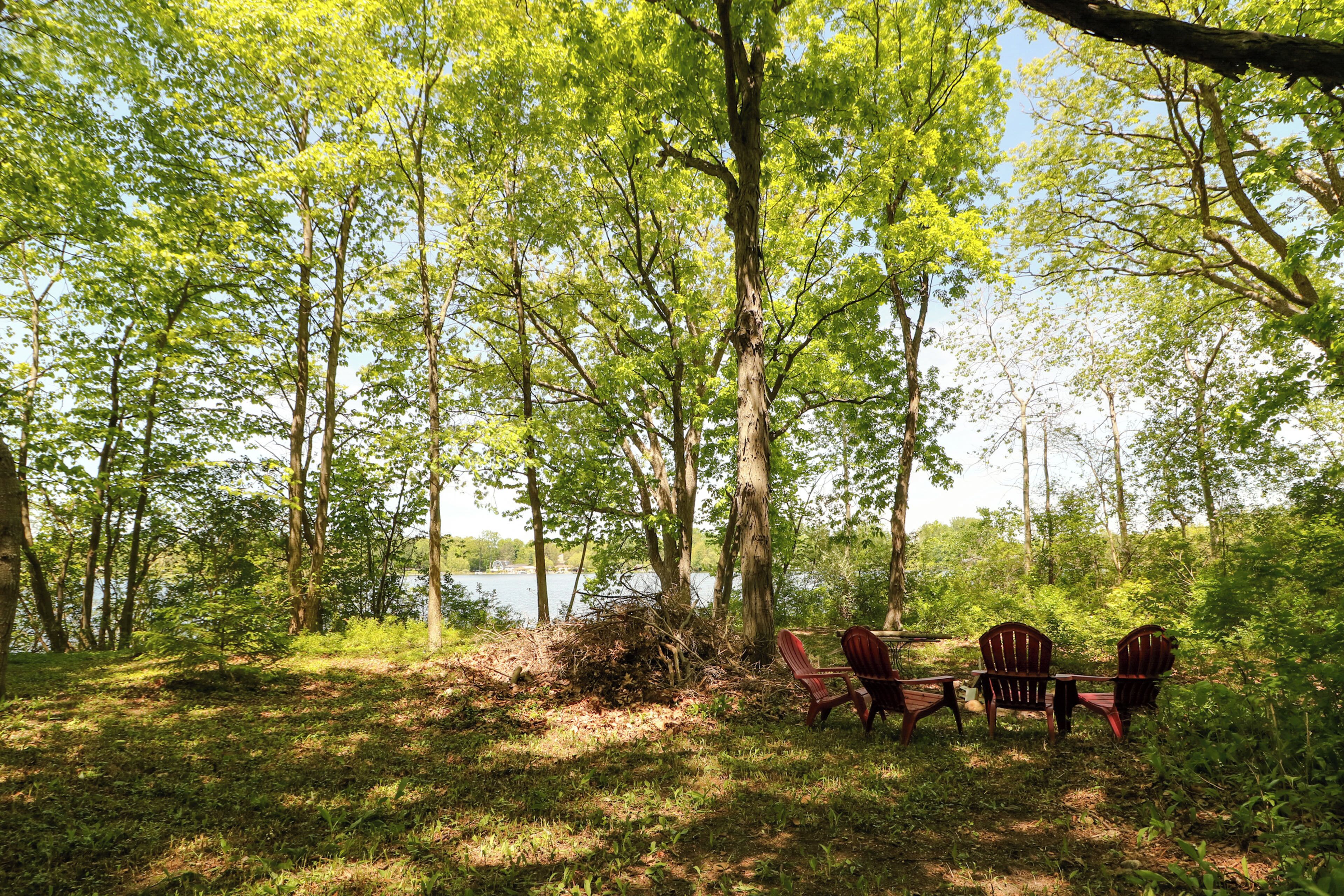 A campsite with a table and four Adirondack chairs near a lake surrounded by trees.