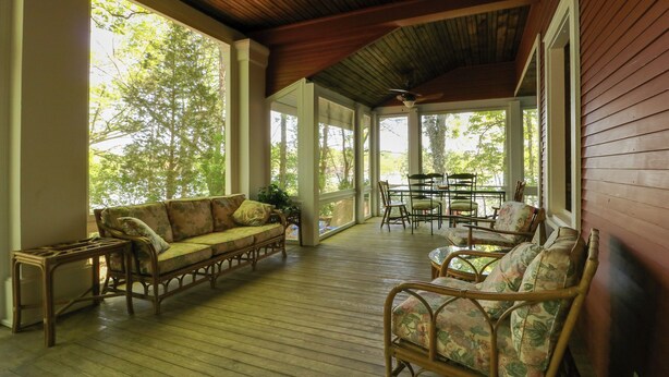 A covered porch with wooden flooring, wicker furniture, and large windows offering views of trees. - Island Cottage on Evans Lake: Boat Rental Required (Tipton)