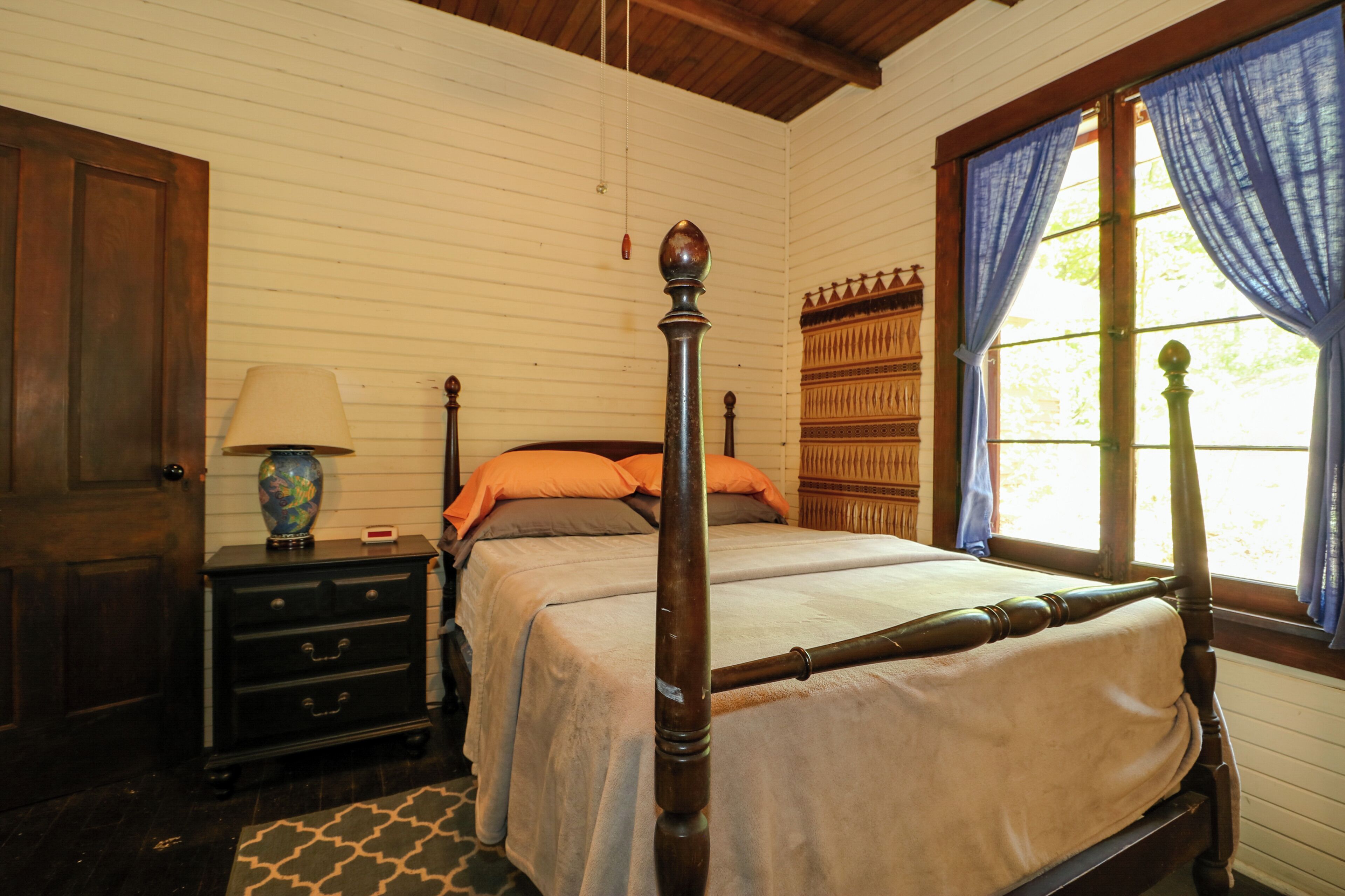 A traditional wooden four-poster bed with a beige bedspread, a nightstand with a lamp, and a patterned rug.