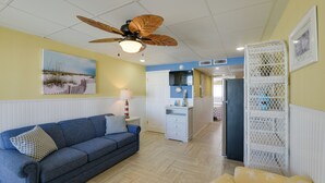 A living room with a blue sofa, a yellow armchair, a ceiling fan, and a kitchen area in the background. - Updated Oceanside Condo - 5 Miles to Cape May! (Wildwood Crest)