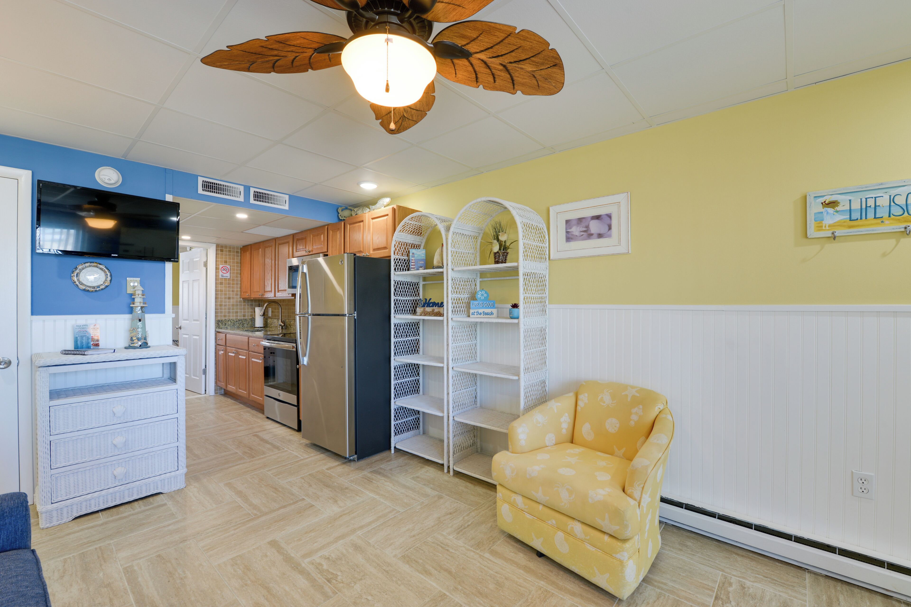 Une cuisine avec un ventilateur de plafond, un fauteuil jaune et un panneau sur lequel on peut lire : « La vie est belle ».