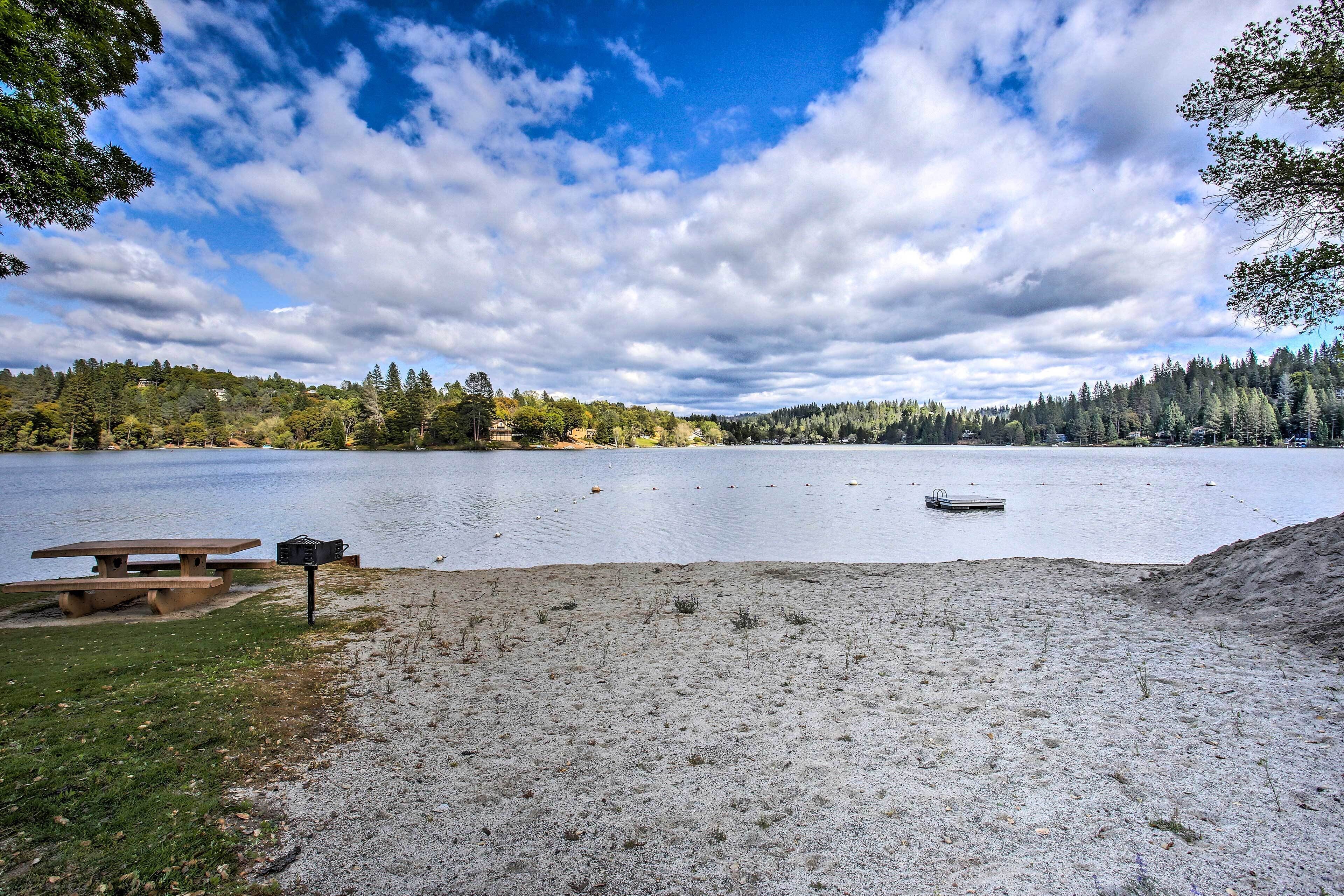 A lakeside picnic area with a bench and a picnic table, surrounded by trees and a clear sky.