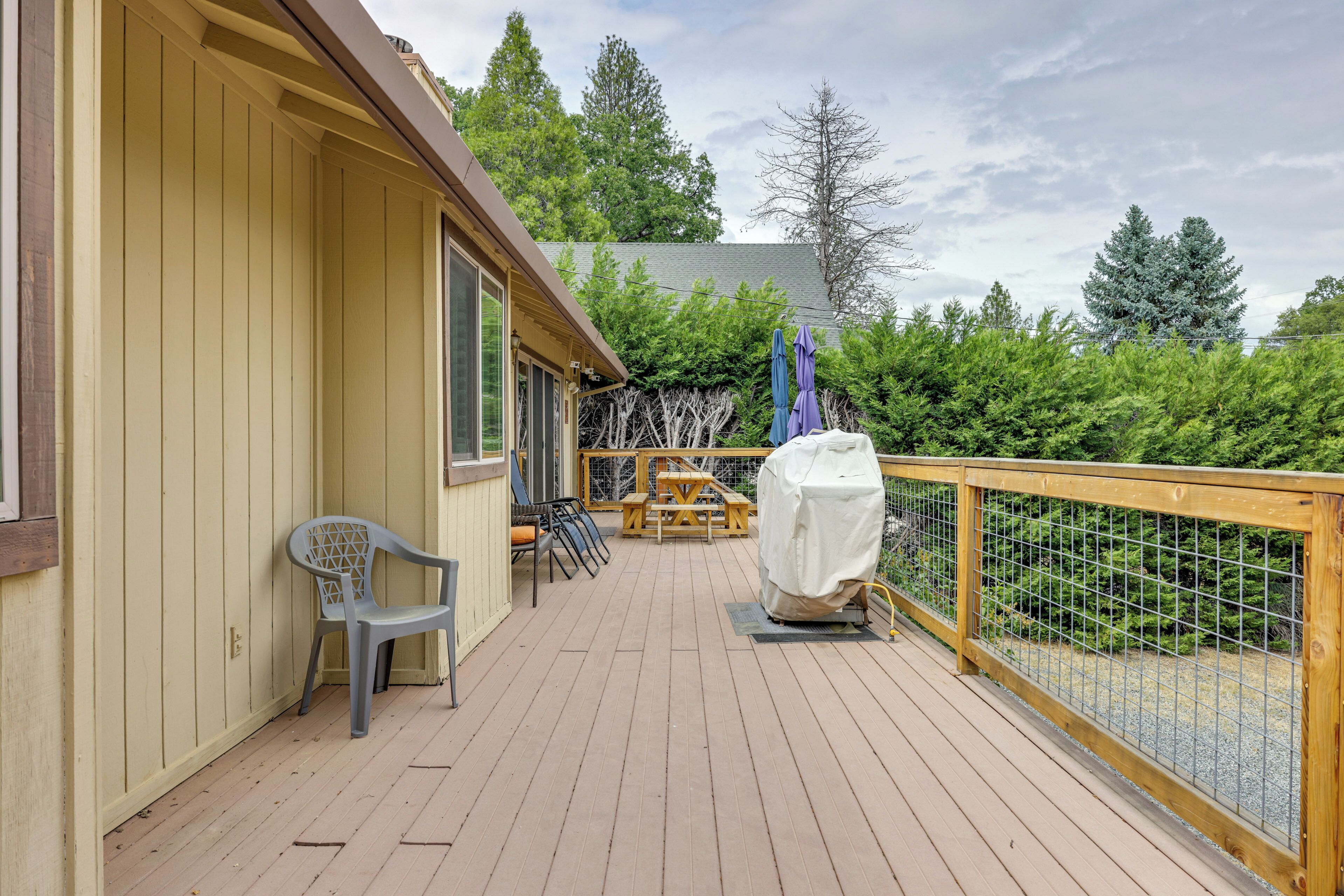 A wooden deck with a covered picnic table and chairs, a covered grill, and a view of the surrounding greenery.
