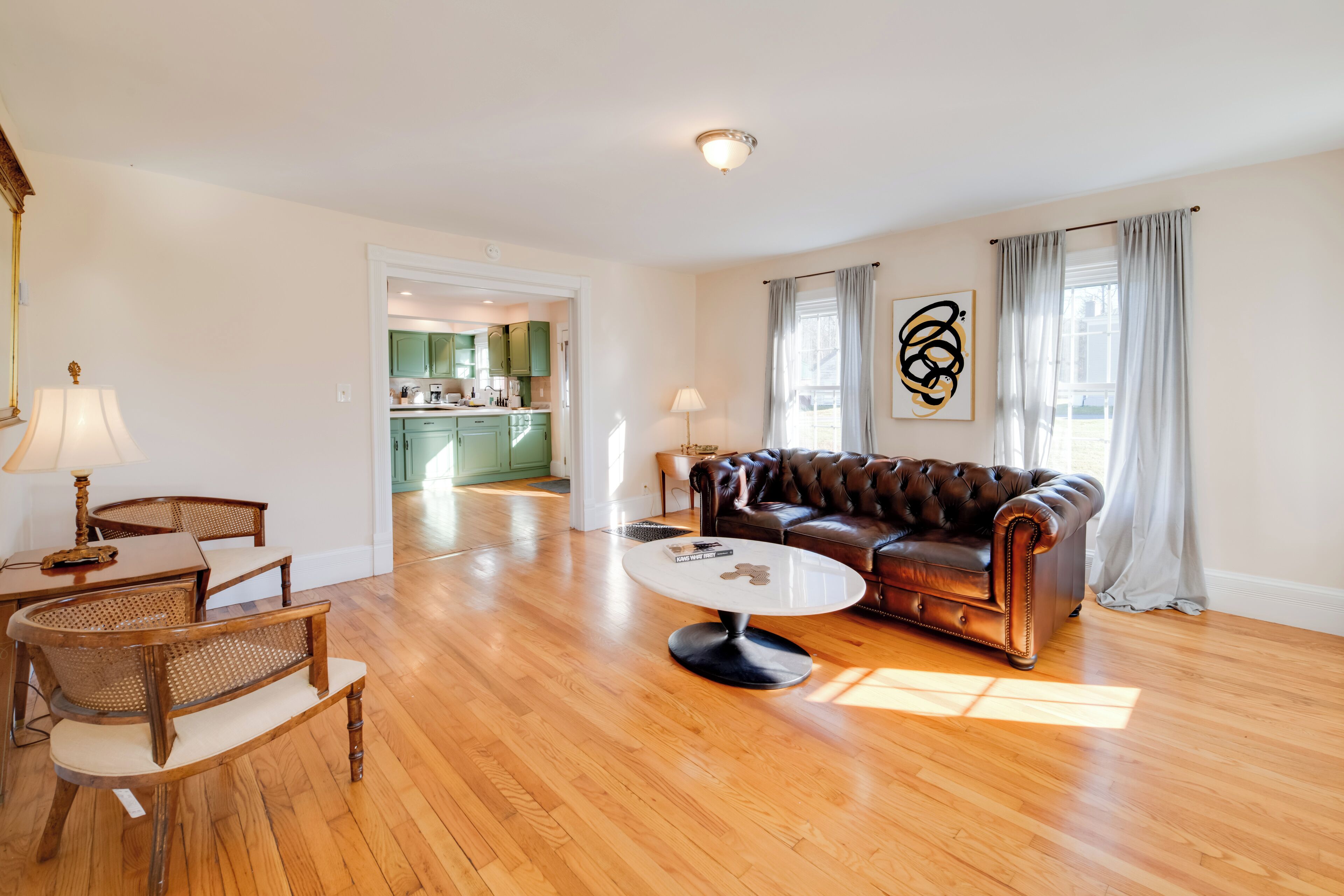 Ein geräumiges Wohnzimmer mit einer Ledersofa, einem Holztisch und Korbstühlen. Im Hintergrund ist eine Küche erkennbar.