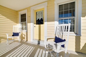 A porch with two white chairs, one with a blue cushion, and a yellow door with a decorative glass panel. - < 4 Mi to Cooperstown: Charming Historic Home! (Fly Creek)