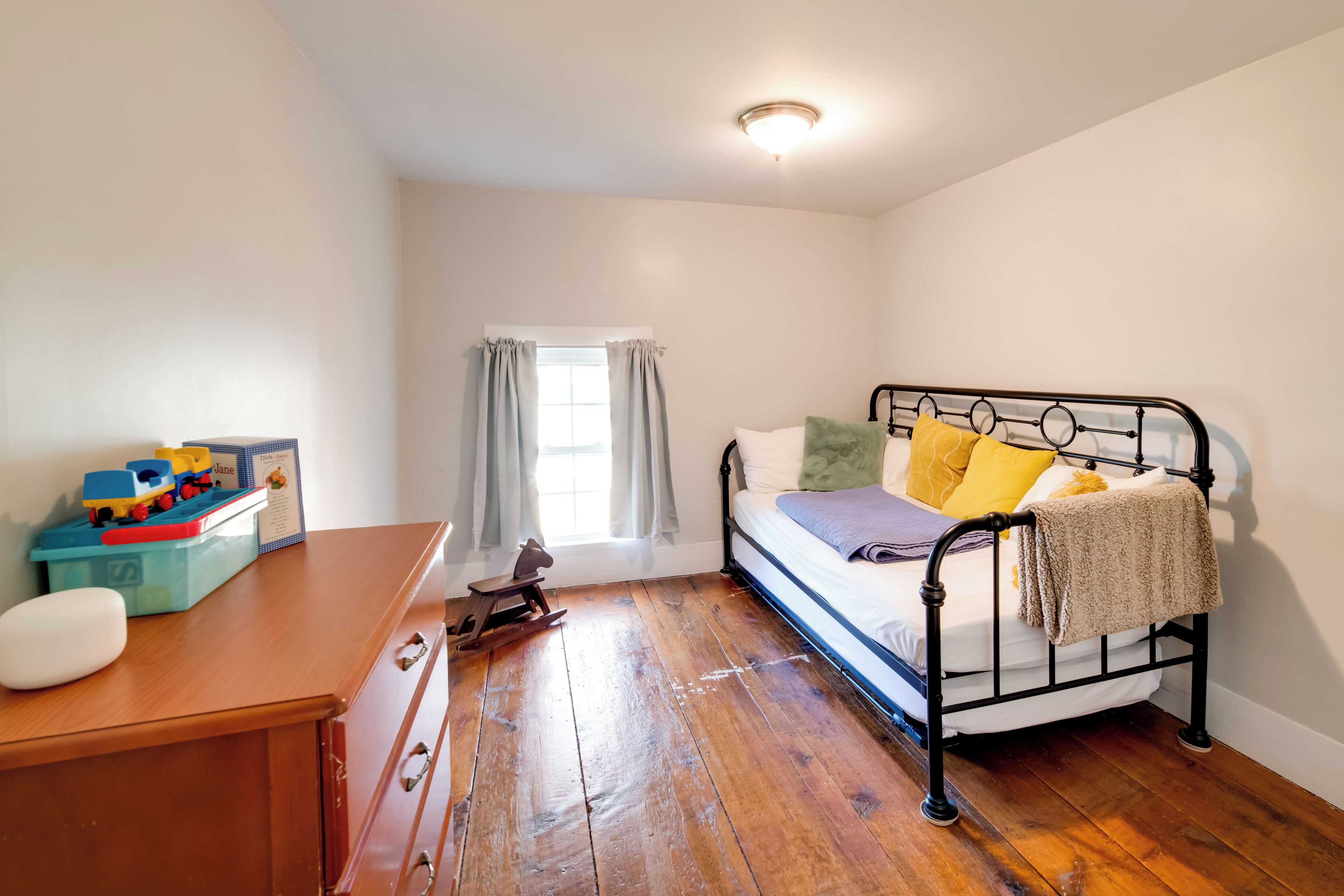 A bedroom with a wooden floor, a bed with pillows, a nightstand, a desk with a chair, and a window with curtains.