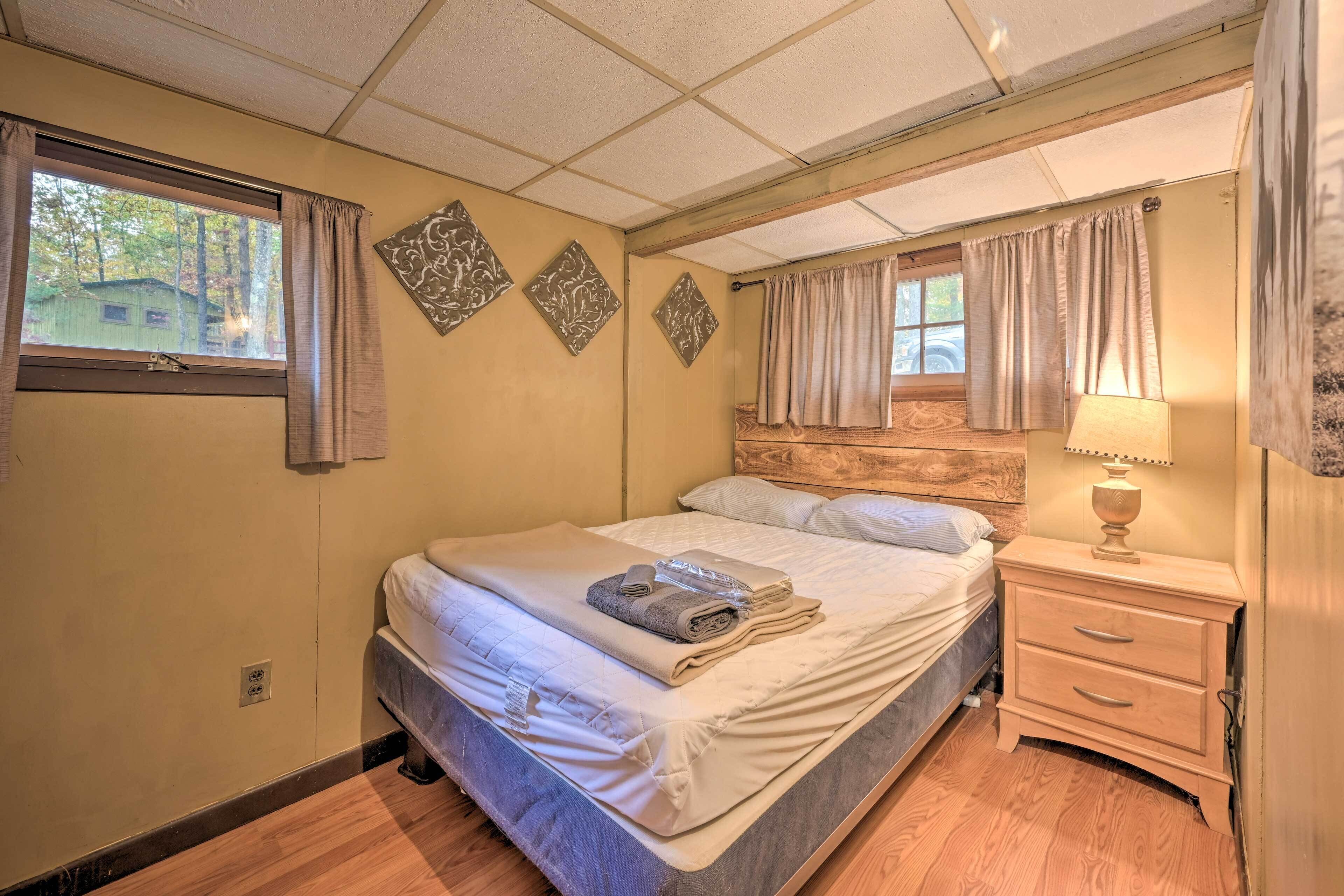 Una cama bien hecha con una toalla doblada y una revista, una mesita de noche con una lámpara, y una ventana con cortinas.