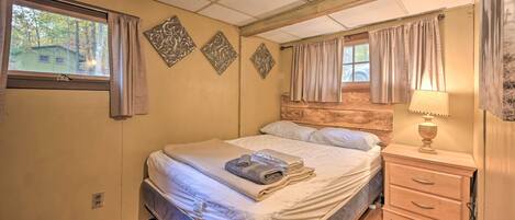 Una cama bien hecha con una toalla doblada y una revista, una mesita de noche con una lámpara, y una ventana con cortinas.