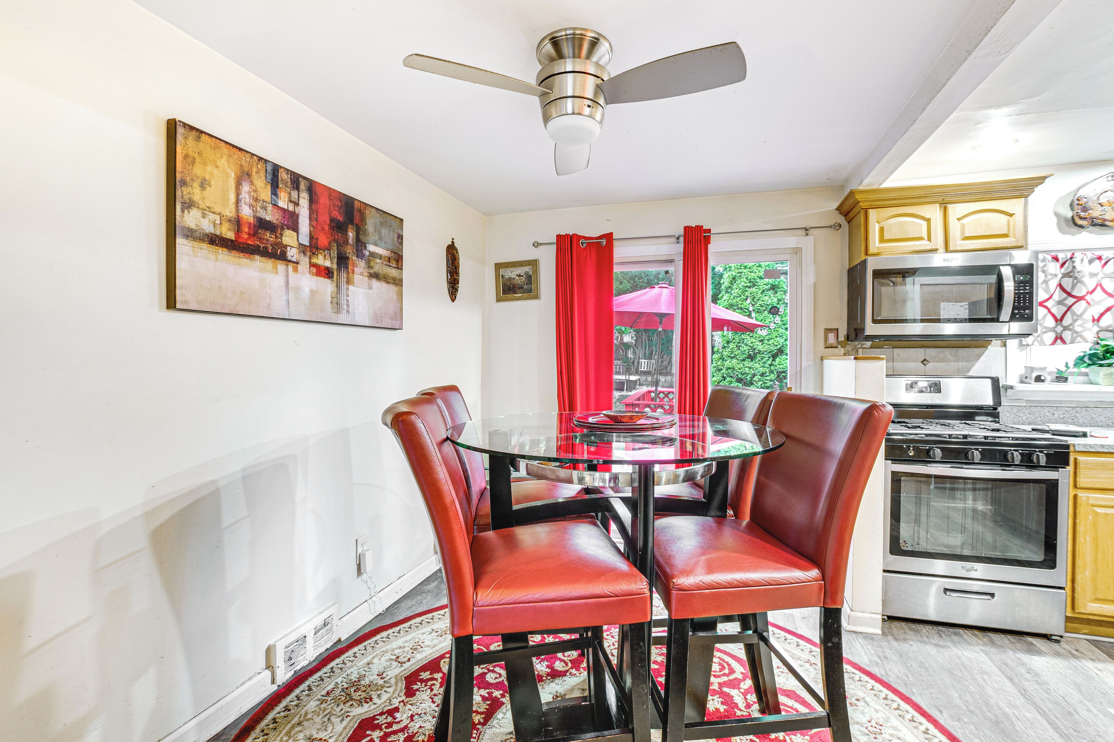 A dining area with a glass table and red chairs, a ceiling fan, a kitchen with modern appliances, and a red curtain.