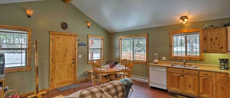 A cozy room with a dining table, chairs, a sofa with a blanket, wooden cabinets, a ceiling fan, and a window with a view of trees.
