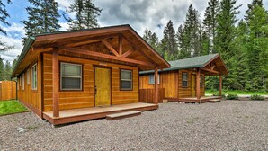A wooden cabin with a porch and a covered area, surrounded by a gravel driveway and dense forest. - Hungry Horse Cabin: Deck, Fire Pit, Near Glacier! (Hungry Horse)