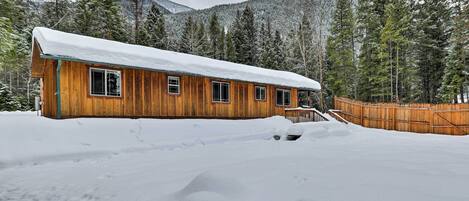 A wooden cabin with a snow-covered roof, surrounded by a wooden fence and snow-covered ground, with a forested mountain backdrop.