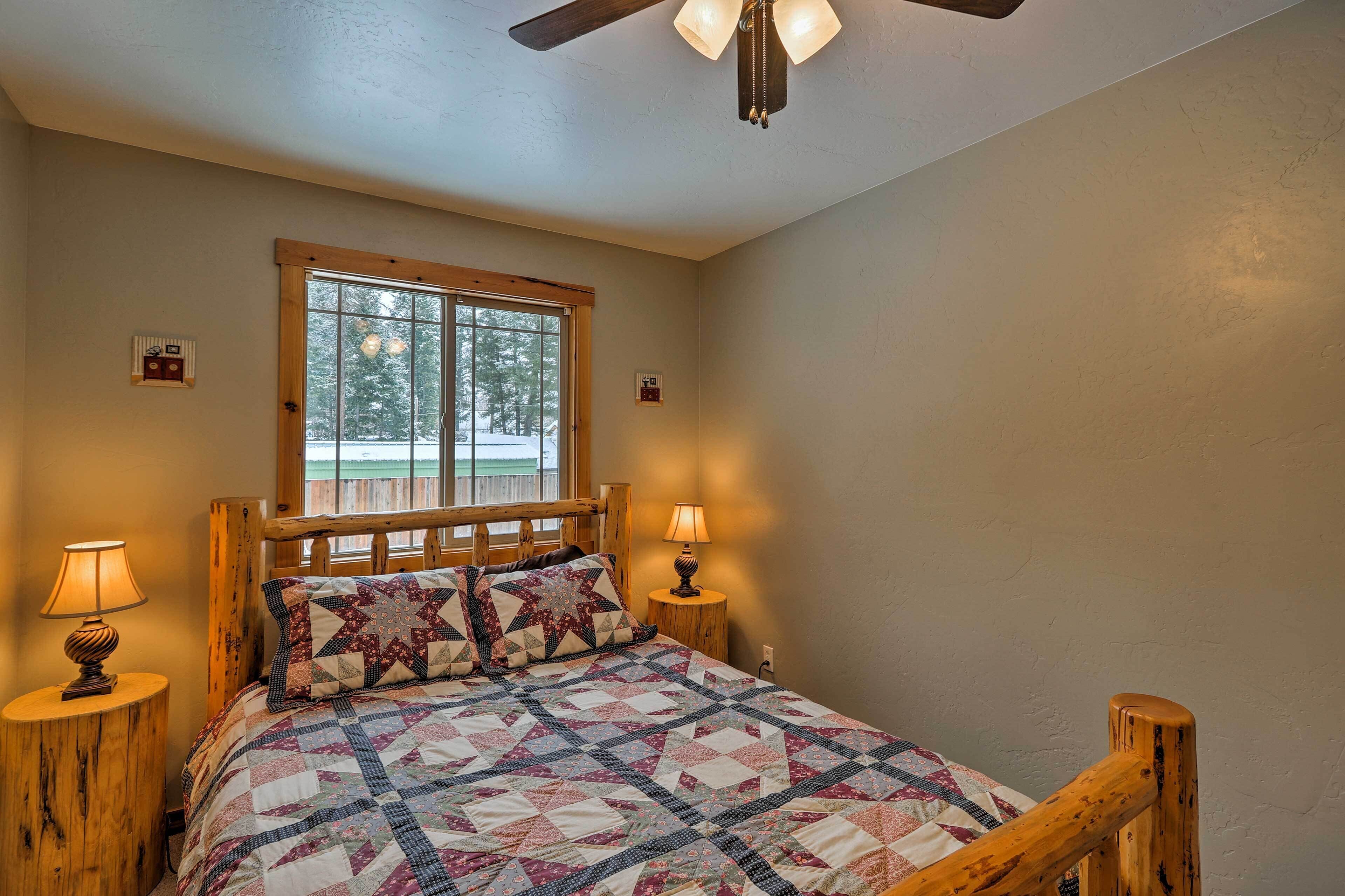 A bedroom with a wooden bed, two bedside lamps, a window with a view of trees, and a ceiling fan.