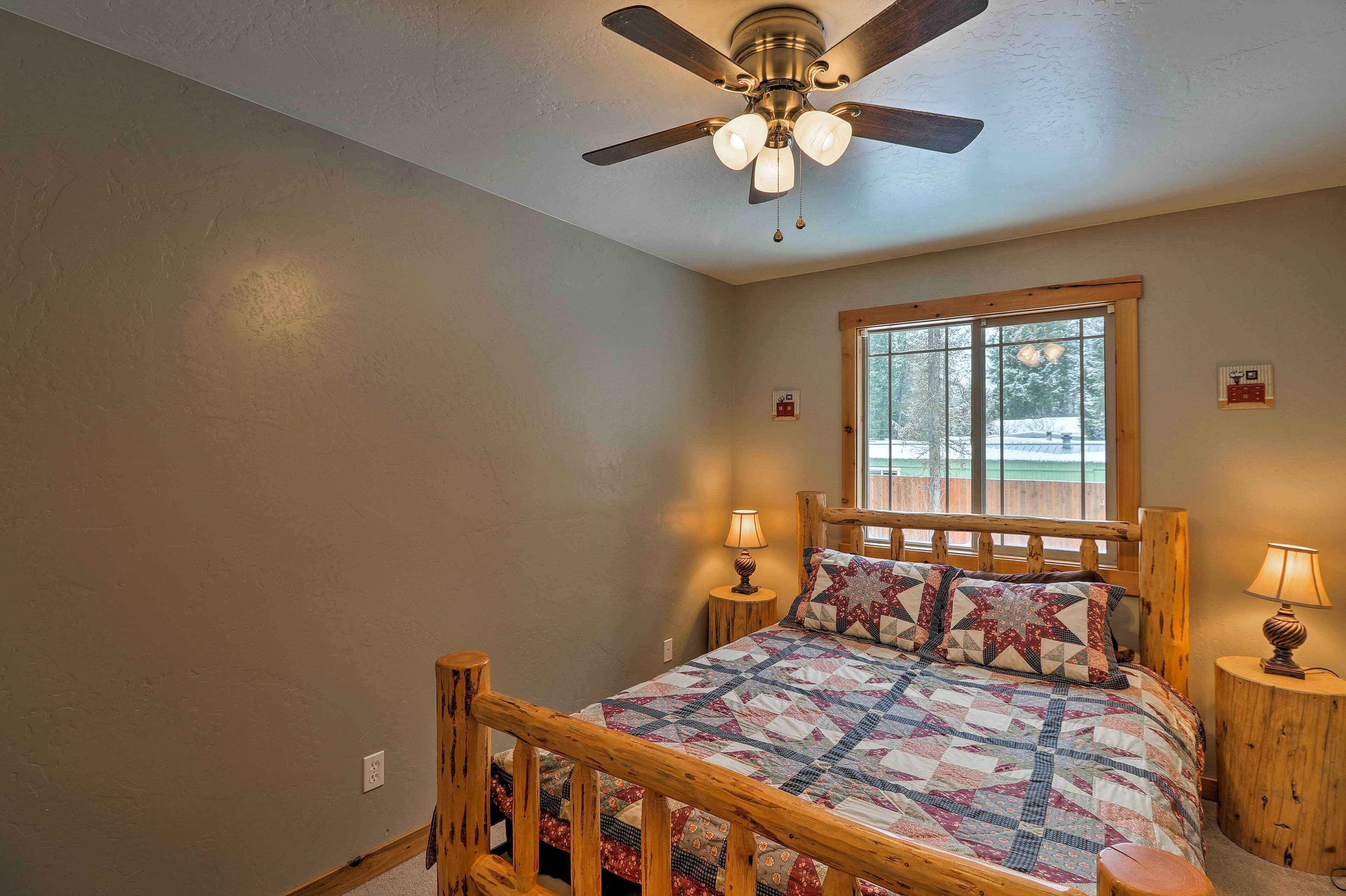 A bedroom with a wooden bed, a ceiling fan, two bedside lamps, and a window with a view of trees.