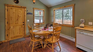 A dining area with a wooden table and chairs, a wooden door, and windows with a view of trees. - Hungry Horse Cabin: Deck, Fire Pit, Near Glacier! (Hungry Horse)