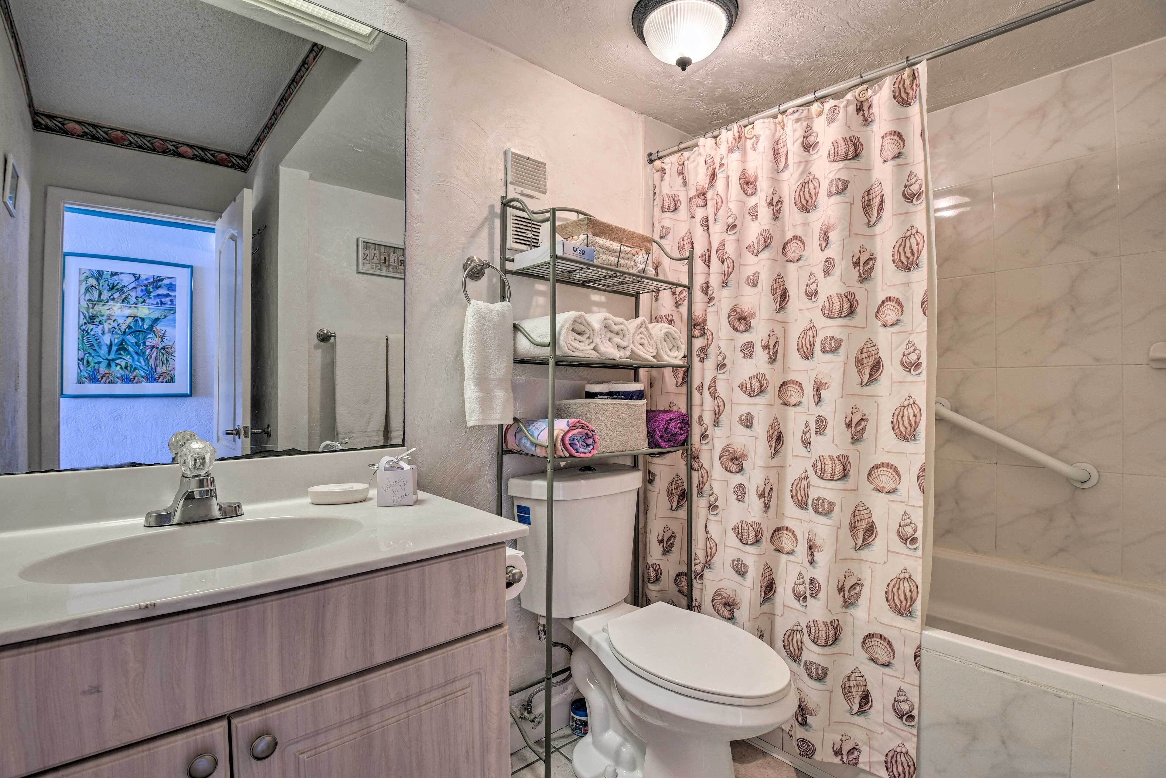 A bathroom with a white sink, toilet, and shower curtain with a shell pattern. There is a towel rack with towels and a shelf with toiletries.