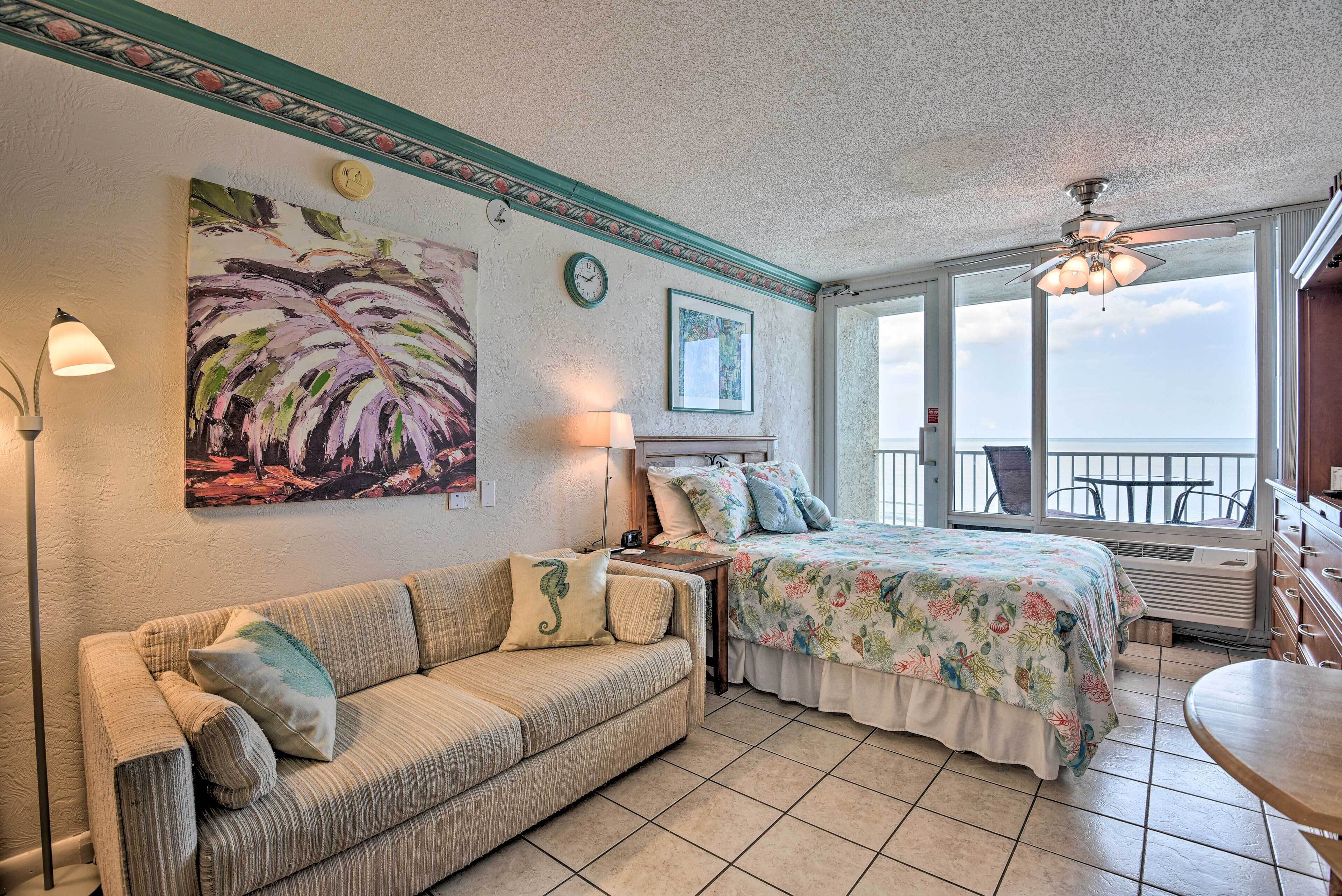 A cozy room with a sofa, a bed with floral bedding, a large painting, a clock, and a view of the outdoors.