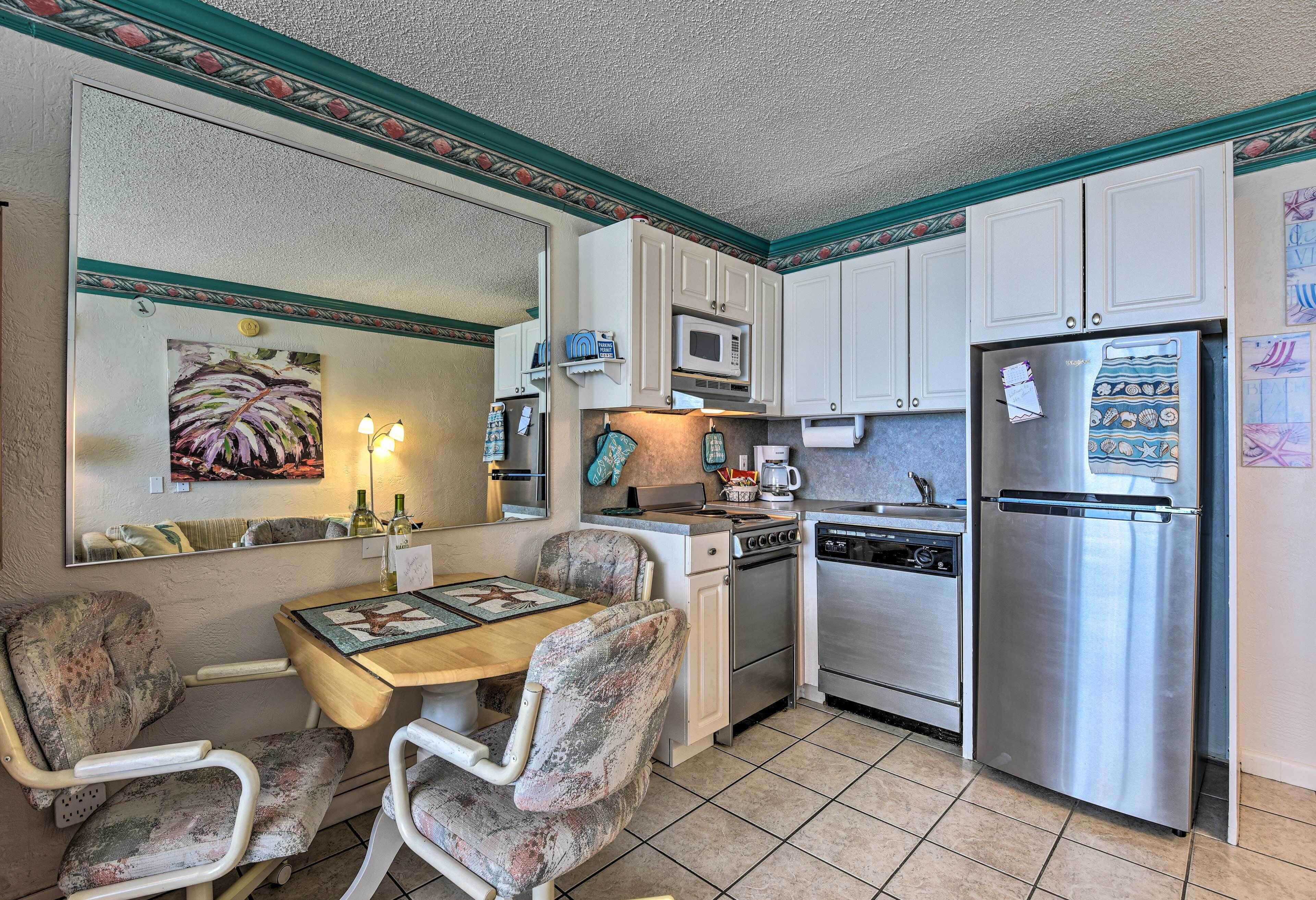 A compact kitchen with white cabinets, a stainless steel refrigerator, a microwave, a stove, and a dishwasher. There is a dining table with two chairs and a painting on the wall.