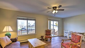 A spacious living room with a wooden bench, two armchairs, a sofa, and a ceiling fan. Large windows provide a view of a deck and outdoor area.