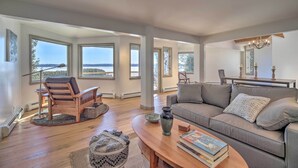 A spacious living room with a wooden floor, a gray sofa, a wooden chair, and a dining table with chairs. Large windows offer a view of the outdoors. - Plattsburgh Home w/ Deck on Lake Champlain (Plattsburgh)