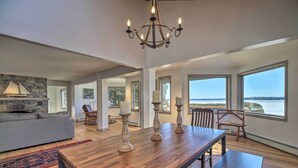 A spacious living area with a wooden dining table, a stone fireplace, and large windows offering a view of the water. - Plattsburgh Home w/ Deck on Lake Champlain (Plattsburgh)