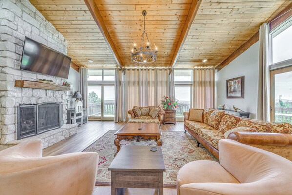 A spacious living room with a stone fireplace, wooden ceiling, and large windows.