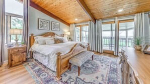 Une chambre spacieuse avec un grand lit, une tête de lit en bois et un banc. On y trouve deux tables de chevet, une commode et une coiffeuse. La chambre est dotée de grandes fenêtres avec des rideaux et offre une vue sur l’extérieur.