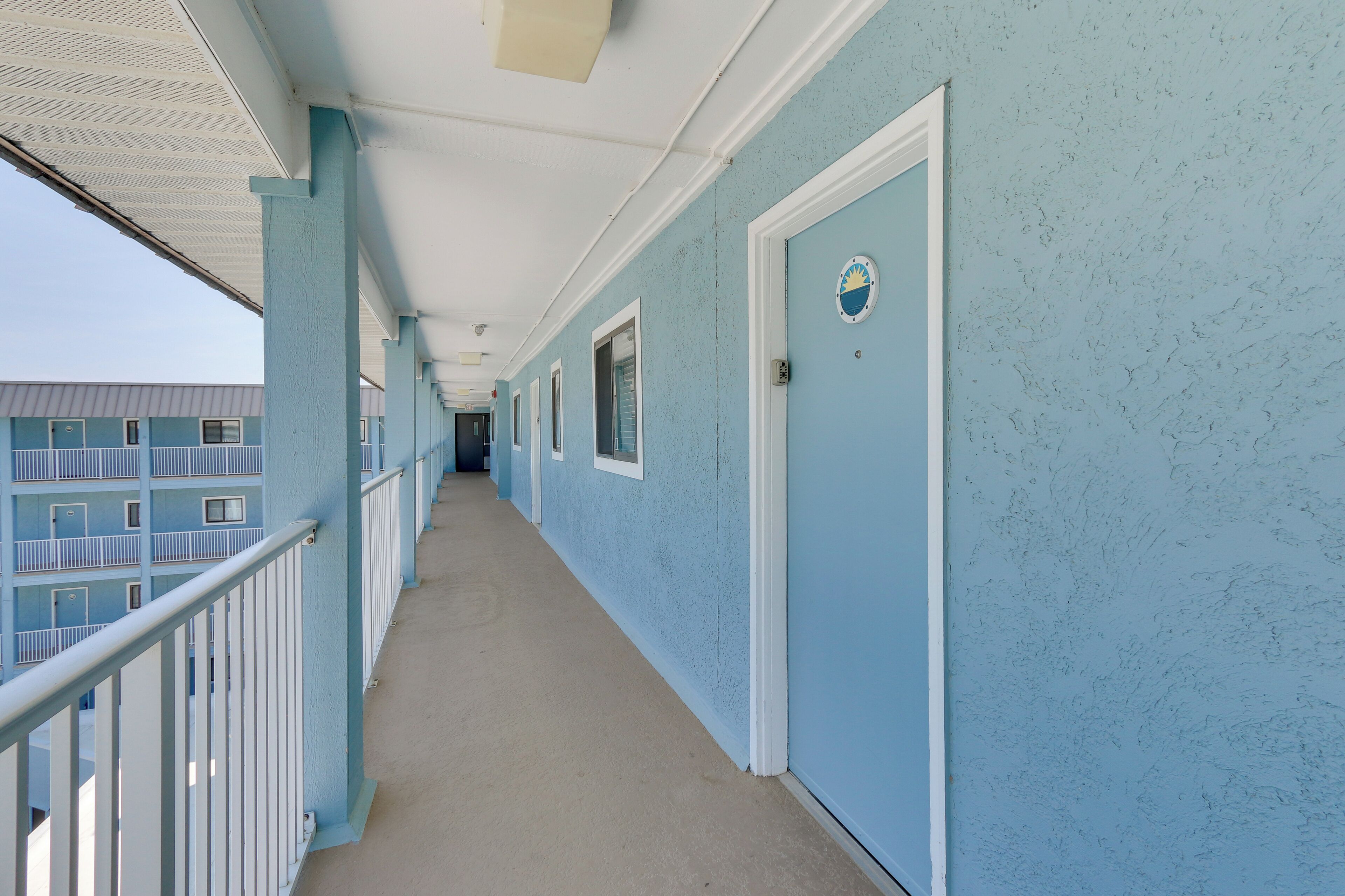 A corridor with light blue walls and white railings, leading to a door with a circular emblem.