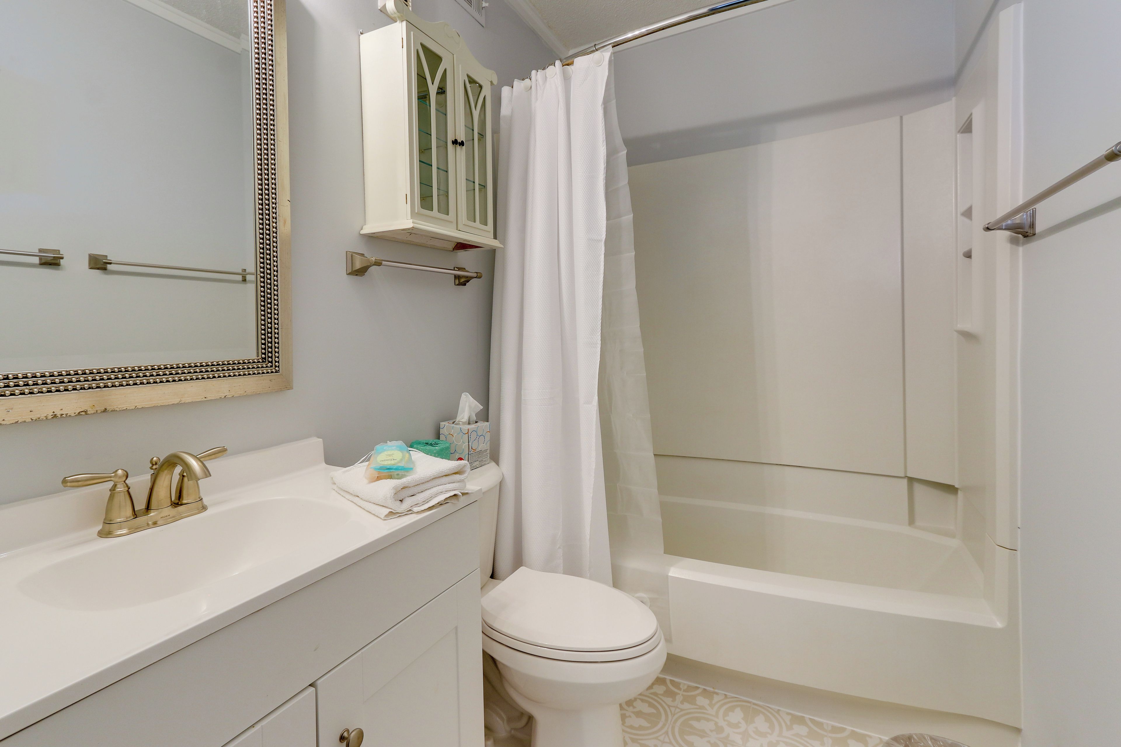 Une salle de bain avec un lavabo blanc, un robinet doré et des toilettes. On y trouve un rideau de douche, un porte-serviettes et un meuble au-dessus du lavabo.