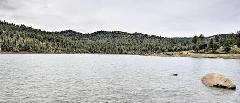 A calm lake with a large rock in the foreground and a forested hill in the background.
