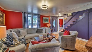 Ein Wohnzimmer mit grauem Sofa, einem Couchtisch und einem grauen Sessel. Eine Treppe mit violettem Wandbelag und eine Tür mit einem Schild sind ebenfalls zu sehen.
