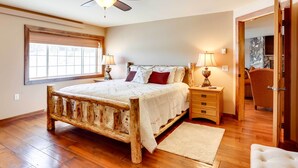 A bedroom with a wooden bed, bedside lamps, a window, and a door leading to another room. - Modern Deadwood Apt With Mountain Views & Grill! (Lead)