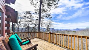 Una terraza de madera con sillas acolchadas verdes, barandilla de madera y vistas a montañas y árboles.