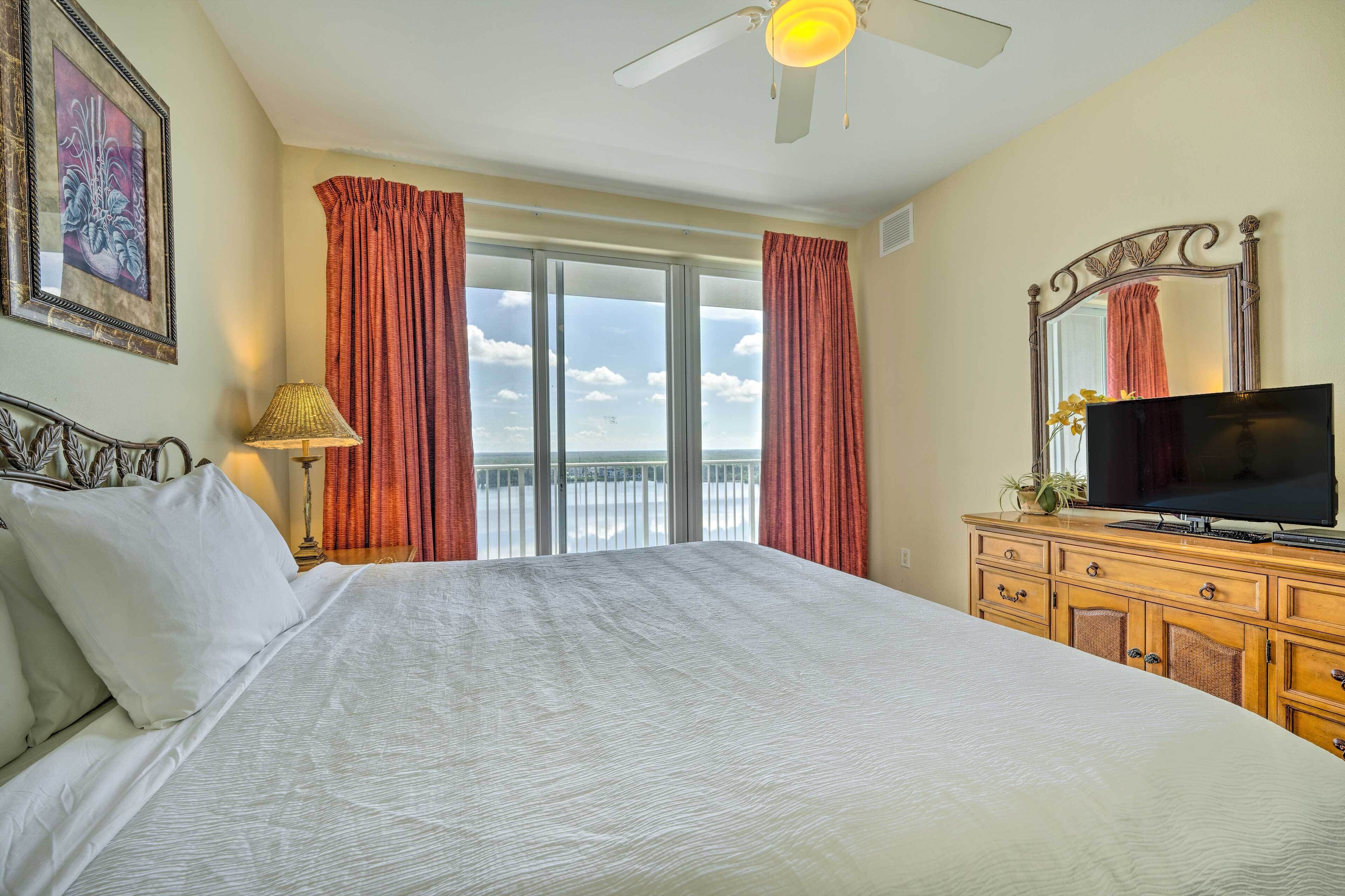 A hotel room with a large bed, a television on a wooden dresser, a ceiling fan, and a balcony with curtains.