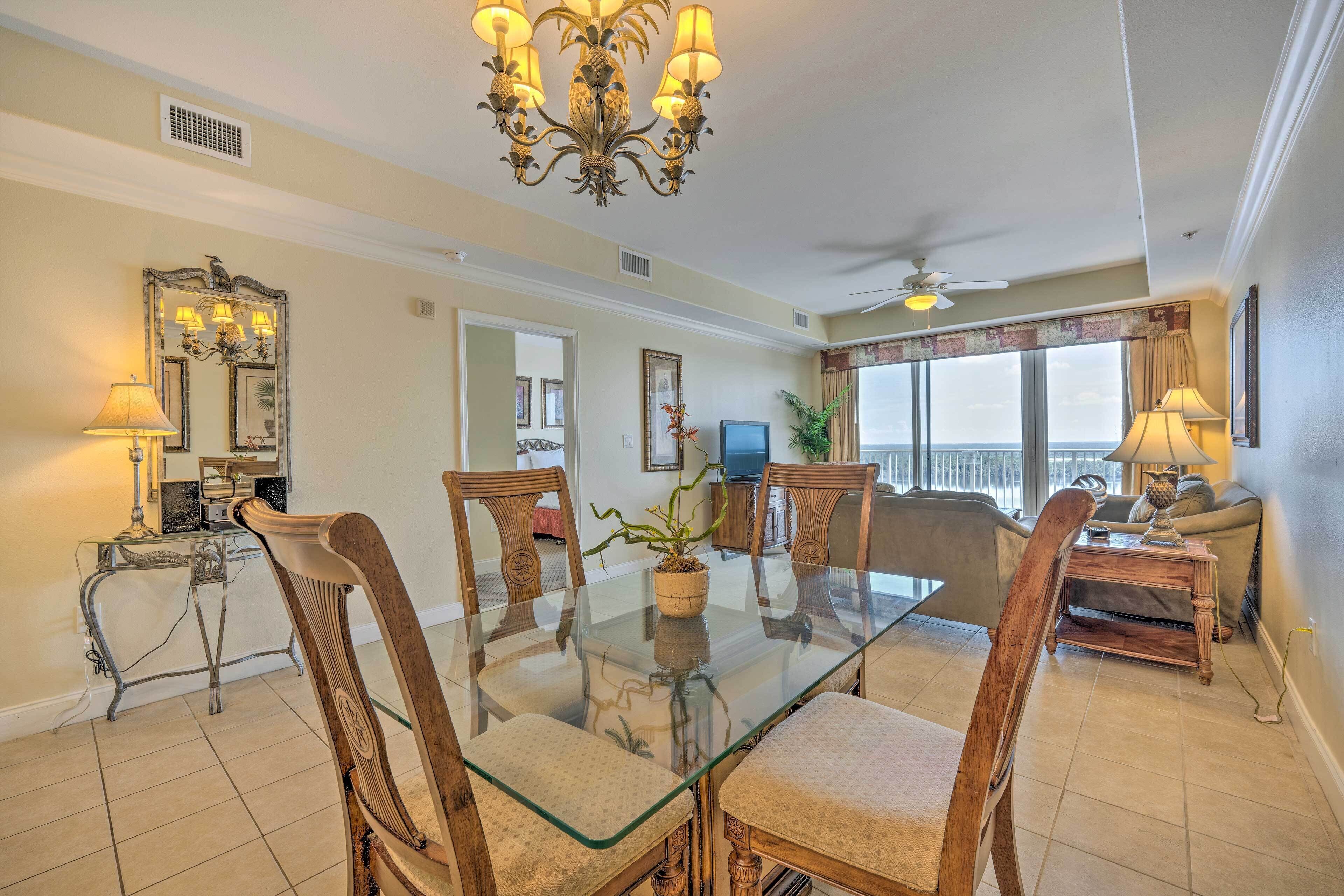 A dining area with a glass table and wooden chairs, a chandelier, and a view of the ocean.